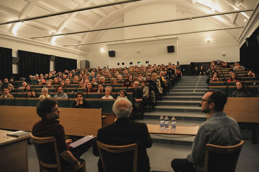 Kolorowa fotografia przedstawia dużą grupę osób w różnym wieku siedzących w dużej sali wykładowej. Słuchają rozmowy po pokazie filmowym. Na pierwszym planie siedzi trzech dyskutujących mężczyzn.