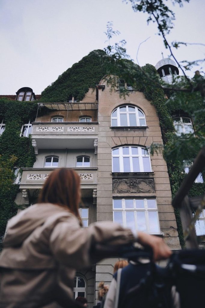 zdjęcie: Anna Pazdej Kolorowa, pionowa fotografia przedstawia starą kamienicę w połowie porośniętą gęstym bluszczem. Pod nią widoczna niewyraźna sylwetka młodej kobiety z rowerem.