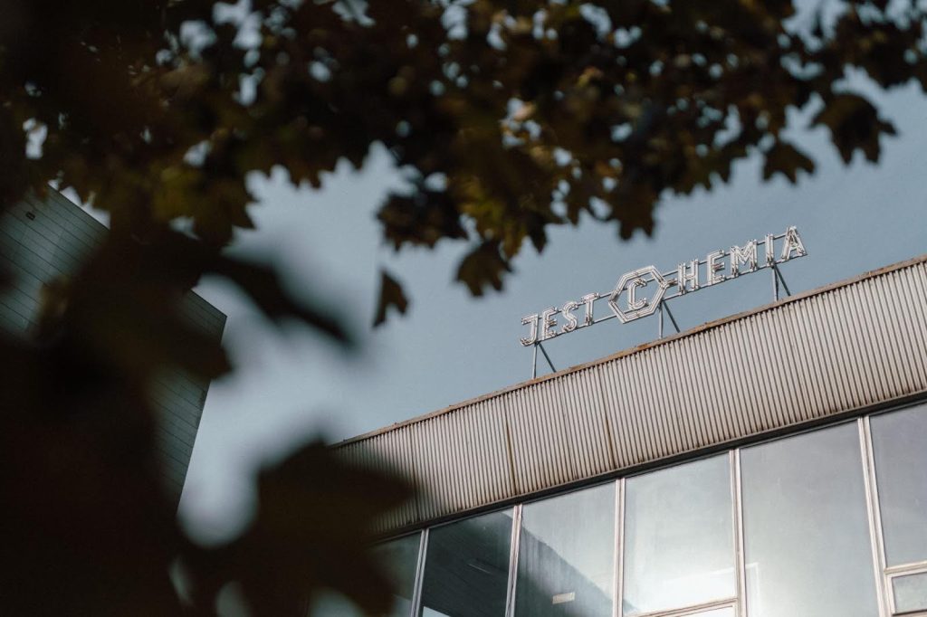 Kolorowa, pozioma fotografia przedstawia neonowy napis "jest chemia" na szczycie budynku.
