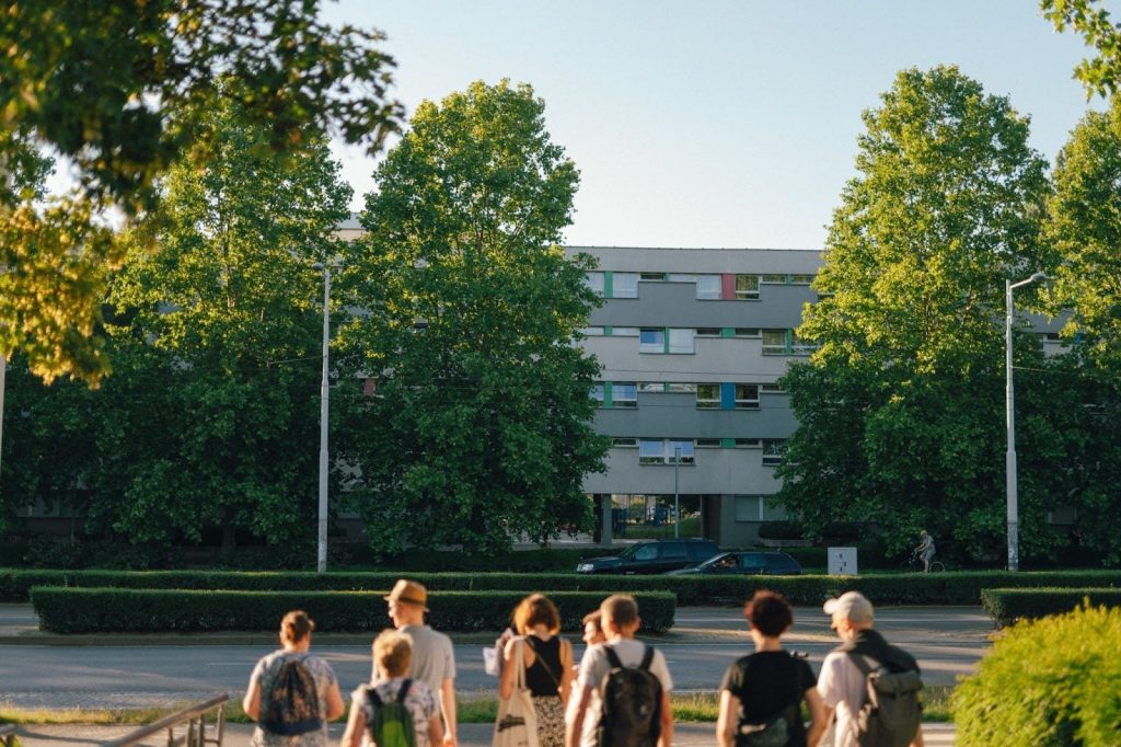 Kolorowa, pozioma fotografia przedstawia grupę osób w różnym wieku spacerujących po osiedlu Plac Grunwaldzki.