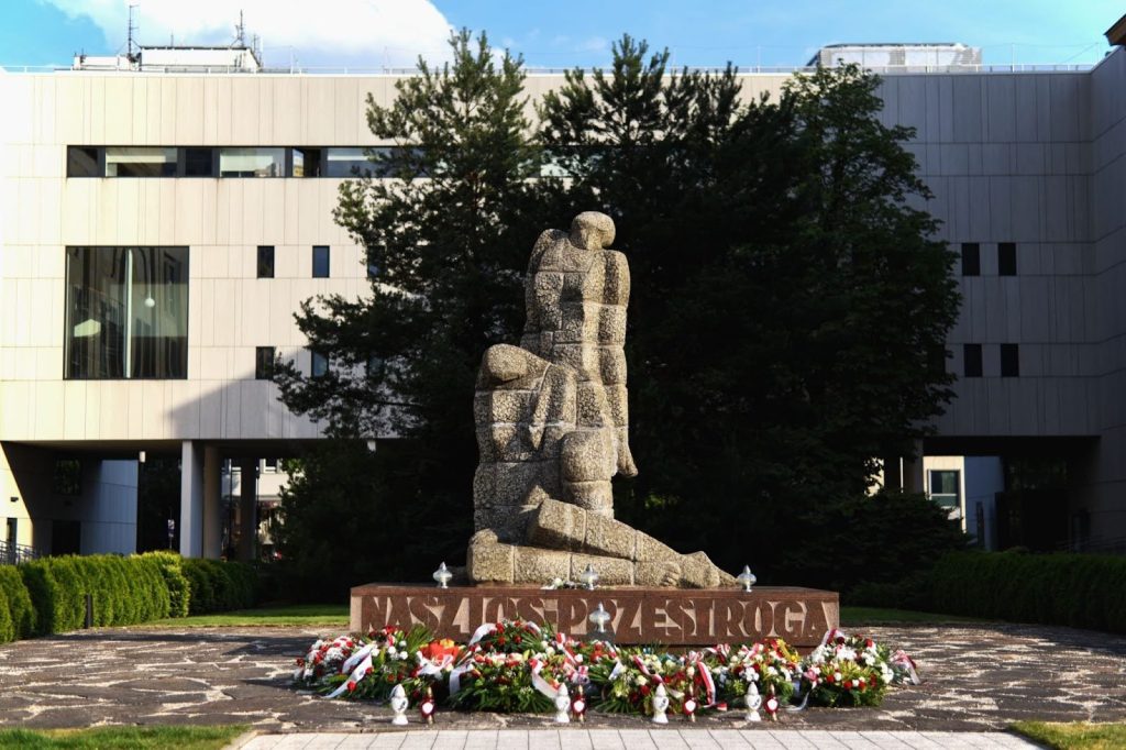 Pozioma, kolorowa fotografia przedstawia widok kamienny pomnik, w tle zabudowa akademicką.