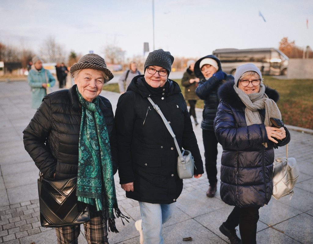 Kolorowa, pozioma fotografia: grupa uśmiechniętych seniorek spaceruje szerokim chodnikiem.