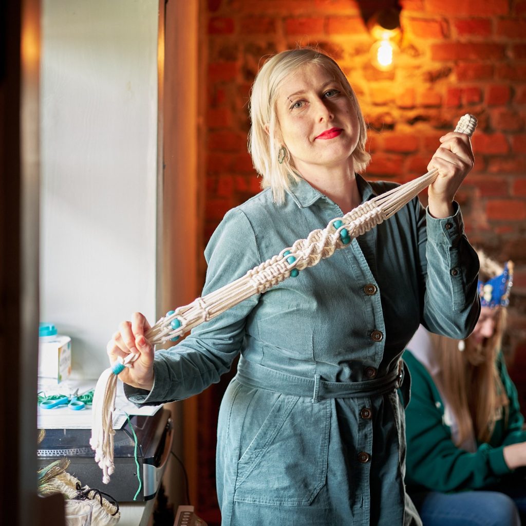 materiały organizatora Kolorowa fotografia: młoda kobieta pozuje do zdjęcia z własnoręcznie plecionym kwietnikiem, ozdobionym niebieskimi koralami. To Monika - prowadząca warsztaty dla seniorów i seniorek.