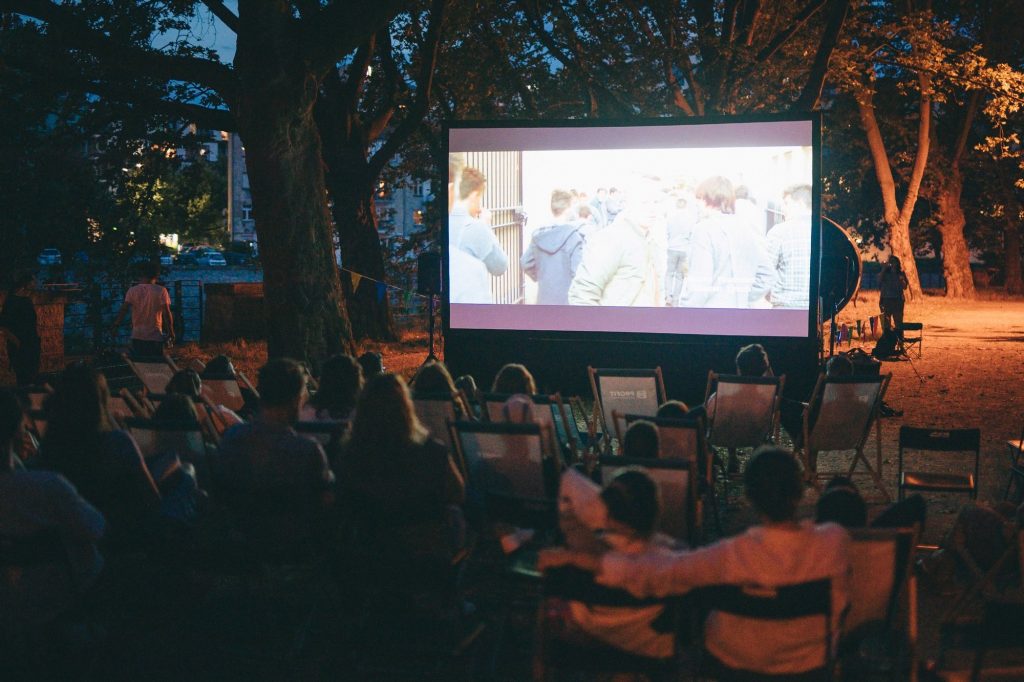 fot. Jerzy Wypych Kolorowa fotografia: duża grupa osób w różnym wieku siedzi na leżakach pod drzewem i ogląda plenerowy pokaz filmu.