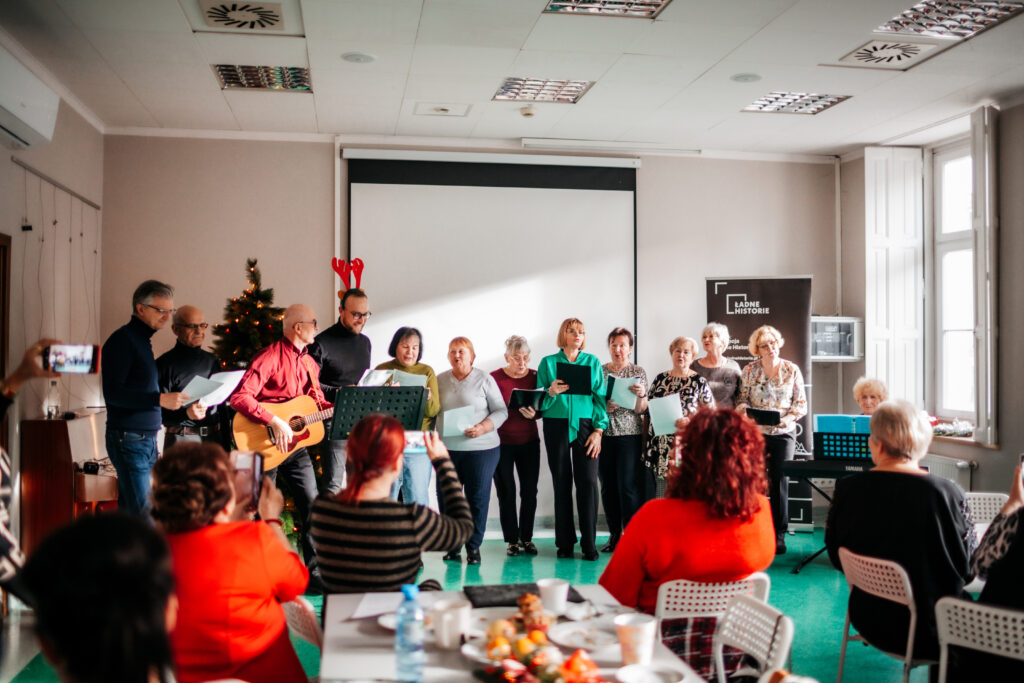 zdjęcie: Kalina Żaczek Kolorowa fotografia przedstawia grupę seniorów i seniorek w jasnym pomieszczeniu. Część z nich stoi na środku i śpiewa, podczas gdy jeden z mężczyzn gra na gitarze, reszta siedzi przy stołach i słucha oraz robi im zdjęcia. Trwa międzykulturowe świąteczne śniadanie.