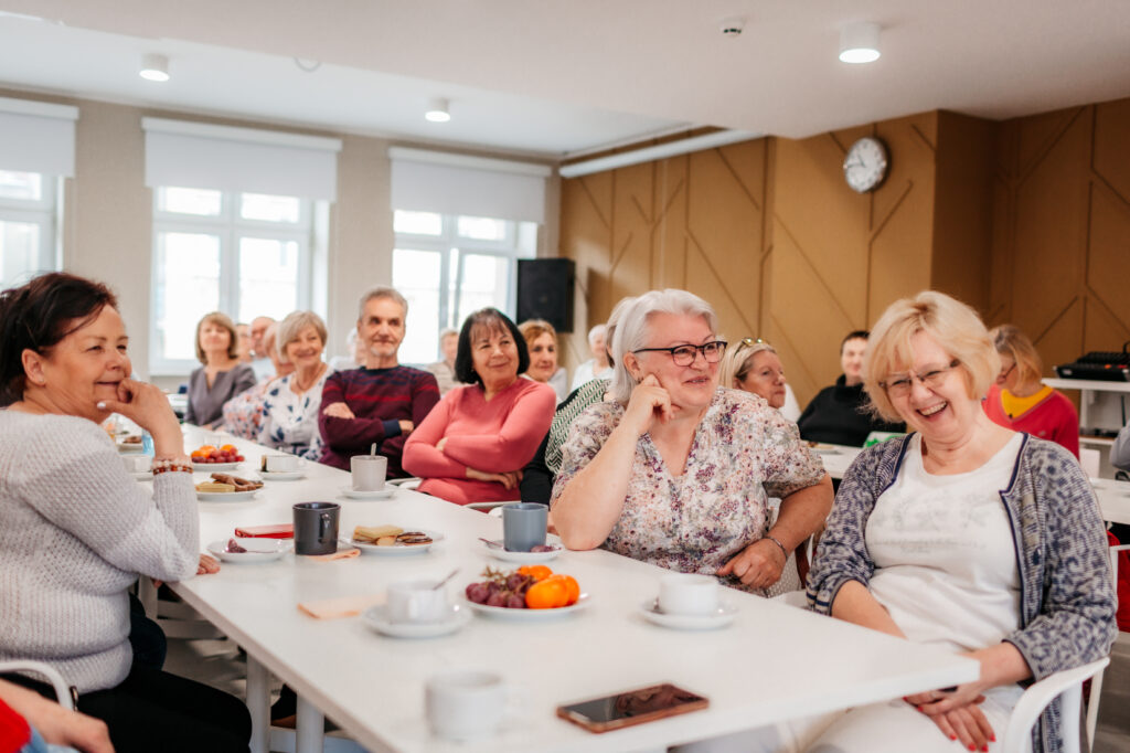 Kolorowa, pozioma fotografia przedstawia grupę seniorów i seniorek siedzących przy długich stołach w jasnym pomieszczeniu. Przed nimi na stole filiżanki z kawą i talerze z owocami i słodyczami. Trwa spotkanie międzykulturowej kawiarenki.