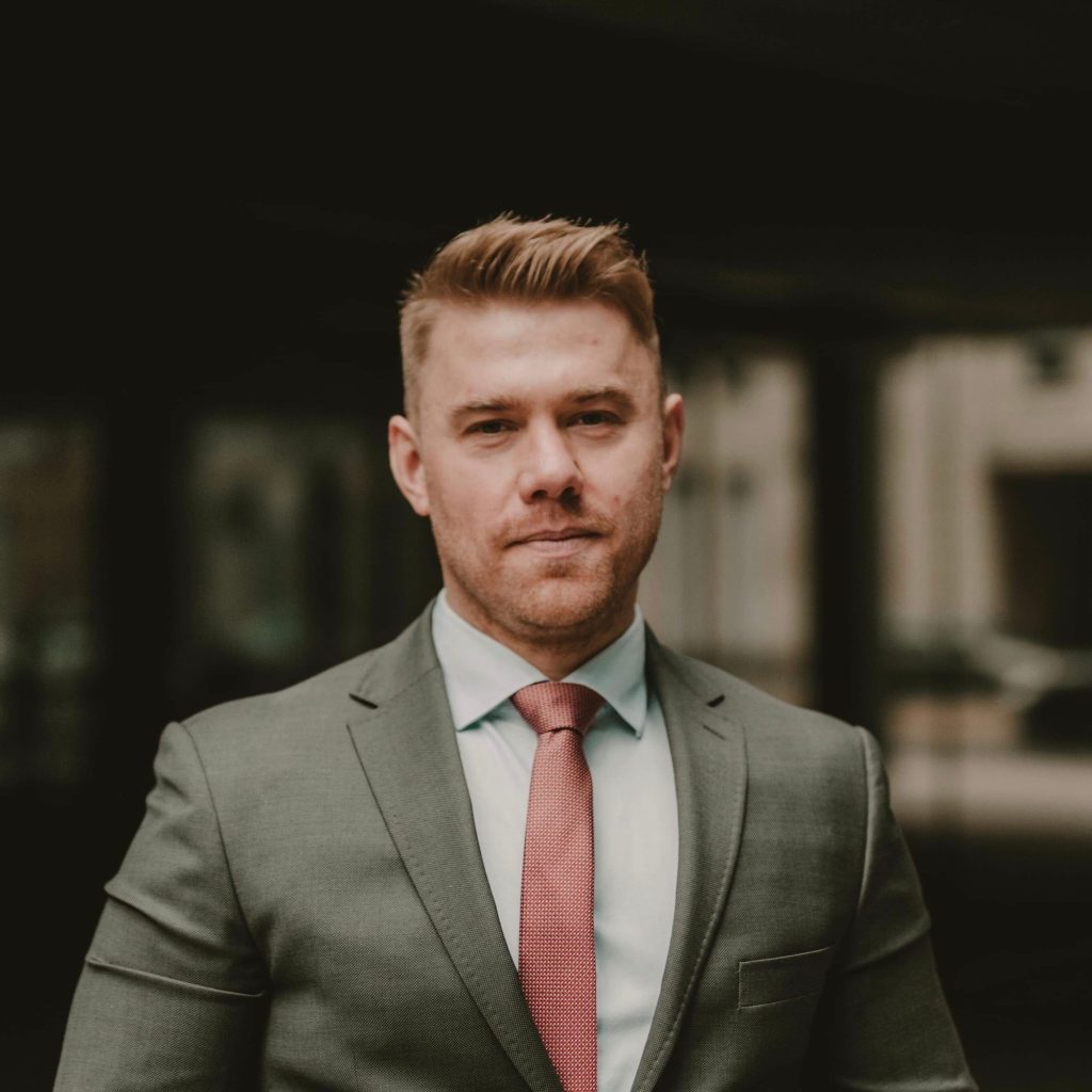 Kolorowa fotografia przedstawia młodego mężczyznę z delikatnym zarostem, ubranego w szary garnitur i różowy krawat.