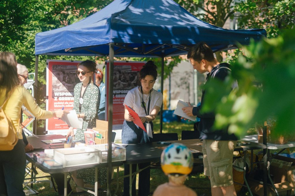 zdjęcie: Marcin Szczygieł Kolorowa, pozioma fotografia wykonana podczas święta osiedla. Pod namiotem rozstawionym na trawie stoją dwie młode dziewczyny i opowiadają o programie wydarzenia osobom stojącym przy stoisku.