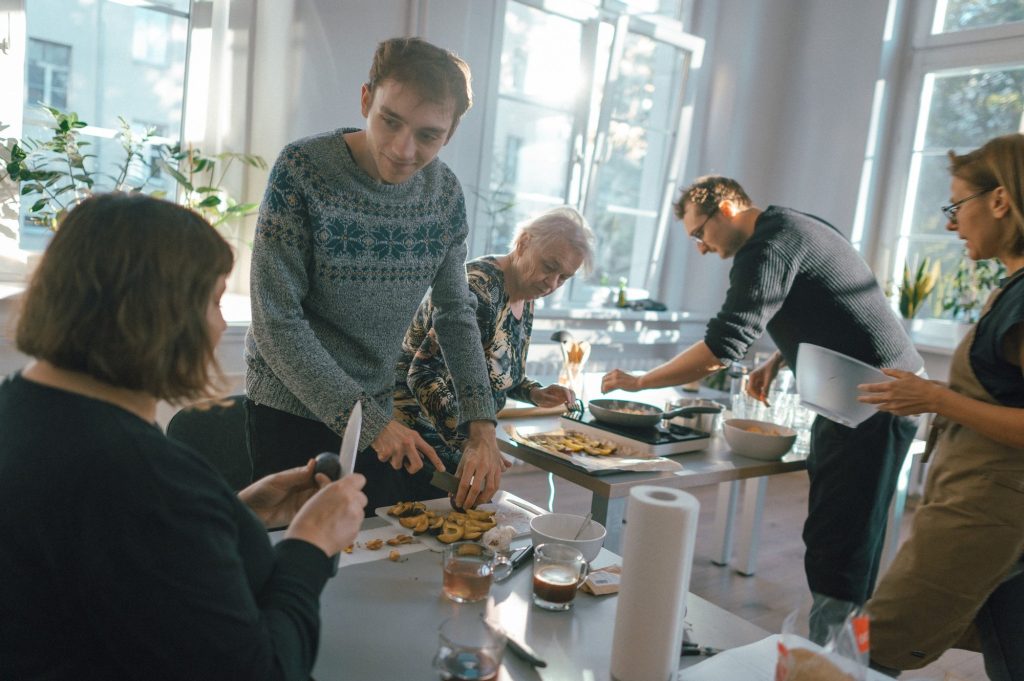 zdjęcie: Marcin Szczygieł Kolorowa fotografia wykonana podczas warsztatów kulinarnych, grupa ośób w różnym wieku przygotowuje potrawy. Dwie osoby kroją owoce, kobieta na drugim planie doprawia przygotowane jedzenie, mężczyzna obok niej smaży coś na patelni.