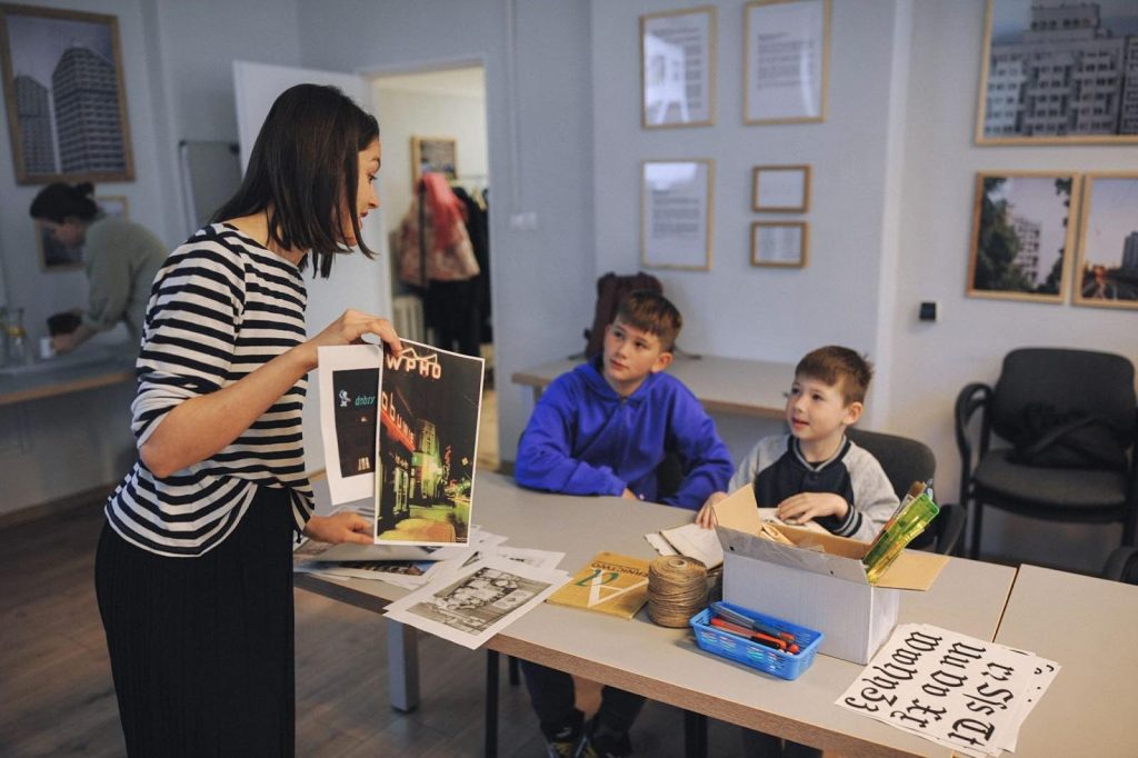Kolorowa fotografia: młoda kobieta prowadząca warsztat pokazuje dwójce młodych chłopców kolorowe grafiki.