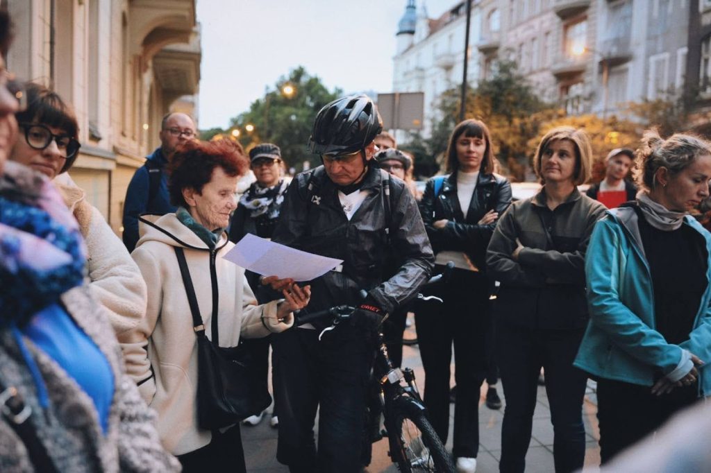 Kolorowa, pozioma fotografia przedstawia grupę ludzi w różnym wieku stojących na ulicy. Trwa spacer po osiedlu.