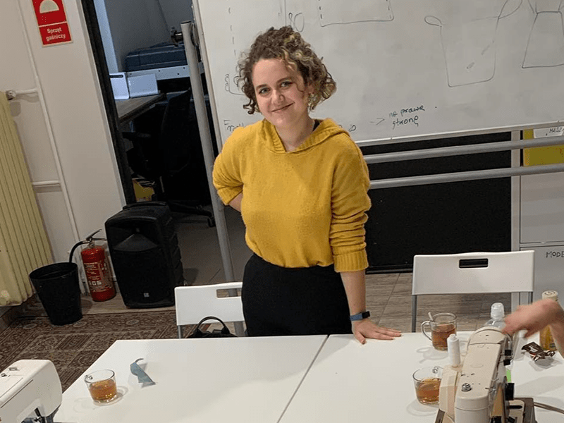 Kolorowa fotografia przedstawia młodą kobietę stojącą przy stole warsztatowym, na którymrozstawione są dwie maszyny do szycia. Kolorowa fotografia przedstawia młodą kobietę stojącą przy stole warsztatowym, na którym rozstawione są dwie maszyny do szycia.