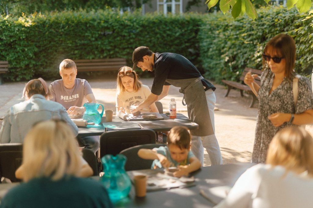 zdjęcie: Marcin Szczygieł Kolorowa fotografia wykonana podczas plenerowych warsztatów ceramicznych. W parku po drzewami rozstawione są stoły, przy których siedzą osoby w różnym wieku i lepią z gliny.