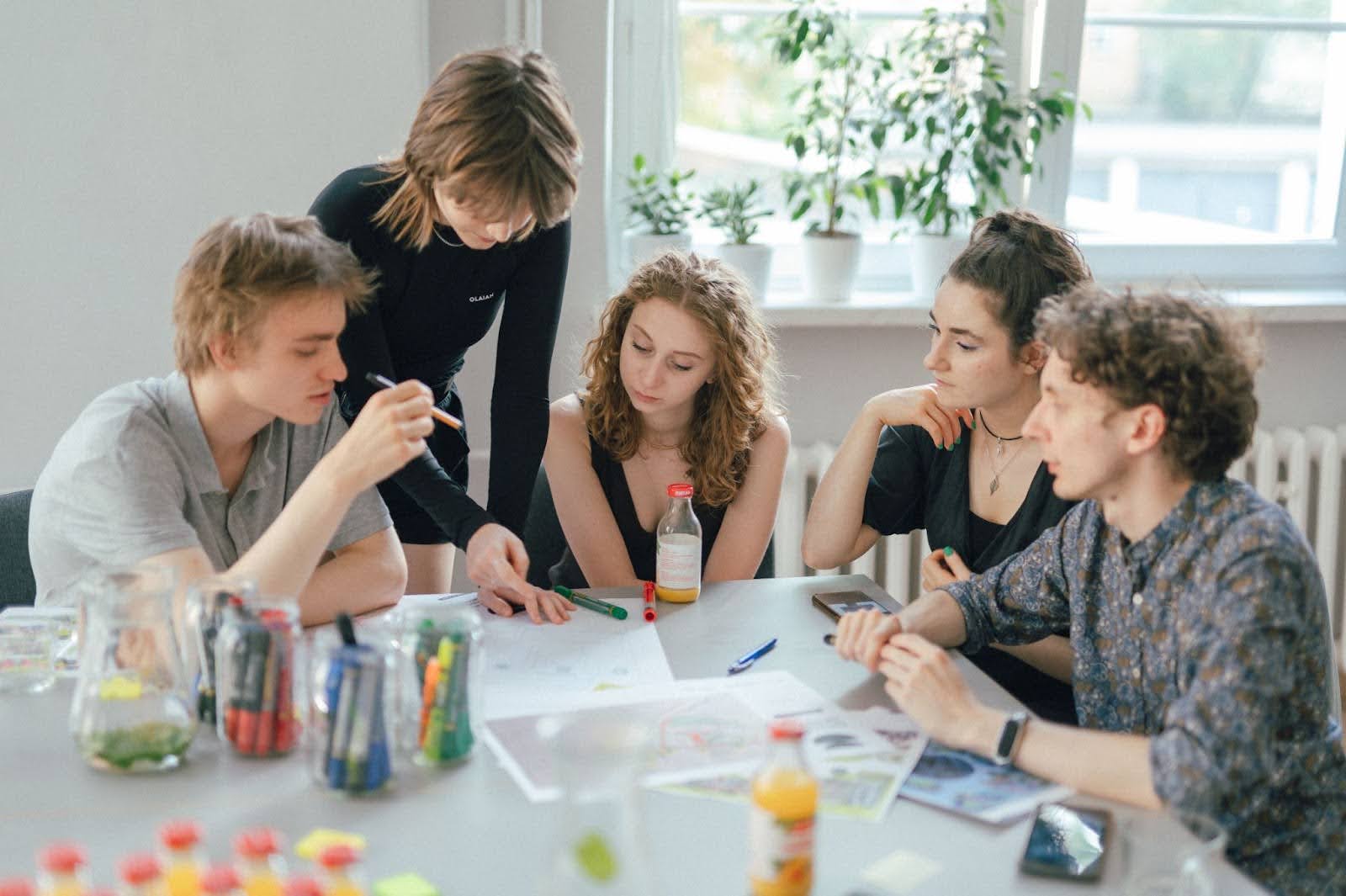 Kolorowa fotografia wykonana podczas warsztatów. W jasnym pomieszczeniu przy stole siedzi grupa osób, jedna z młodych kobiet stoi. Osoby rozmawiają ze sobą, wpatrując się w leżące przed nimi na stole dokumenty. Na stole leżą także mapy oraz różne długopisu i mazaki.