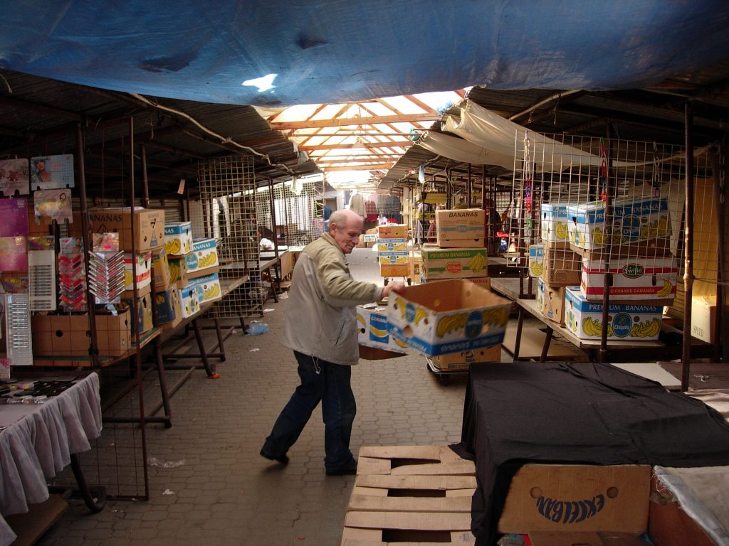 Kolorowa archiwalna fotografia przedstawia targowisko na placu Grunwaldzkim. Na pierwszym planie widoczny mężczyzna przenoszący kartony między straganami.