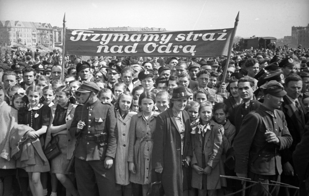 Archiwalne, czarno-białe zdjęcie. Manifestacja na placu Grunwaldzkim w latach czterdziestych XX wieku – w tle kamienice przy ul. Grunwaldzkiej. Na pierwszym planie młodzież trzymająca transparent z napisem: trzymamy straż nad Odrą. Przed nimi stoją żółnierze.