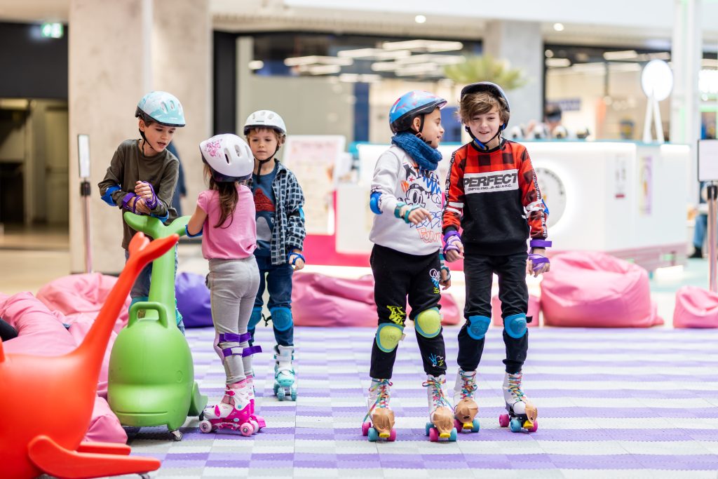 zdjęcie: Pasaż Grunwaldzki Kolorowa fotografia: w przestrzeni centrum handlowego trwają warsztaty dla dzieci z jazdy na wrotkach. Na zdjęciu pięcioro dzieci jeździ w kaskach i ochraniaczach.