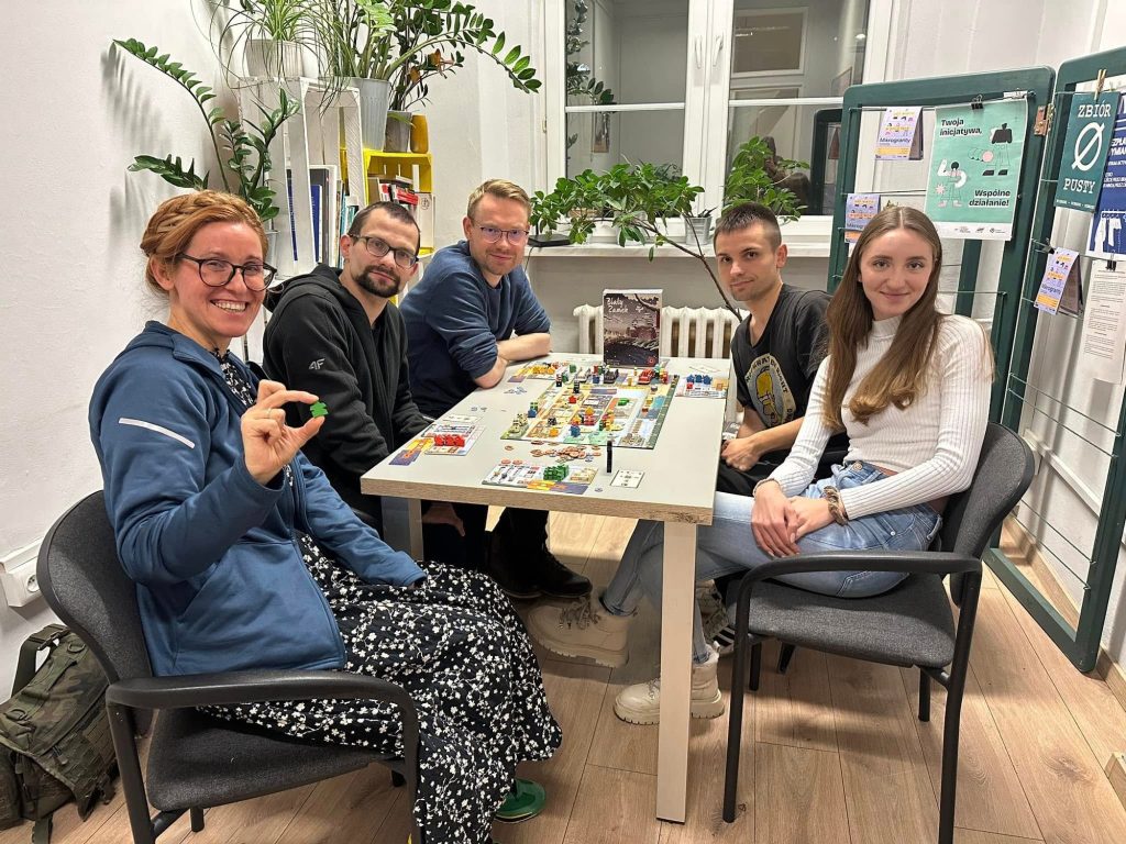 Kolorowa fotografia: w jasnym pomieszczeniu przy stole siedzą osoby w różnym wieku i grają w grę planszową. Trwa spotkanie Grunwaldzkiego Klubu Planszowego.