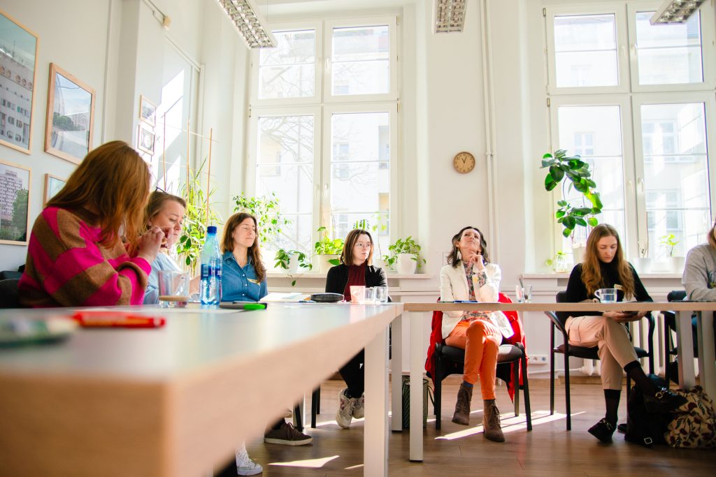 Kolorowa fotografia wykonana podczas warsztatu antywypaleniowego. W dużej jasnej sali, przy długich stołach siedzi grupa kobiet w różnym wieku.