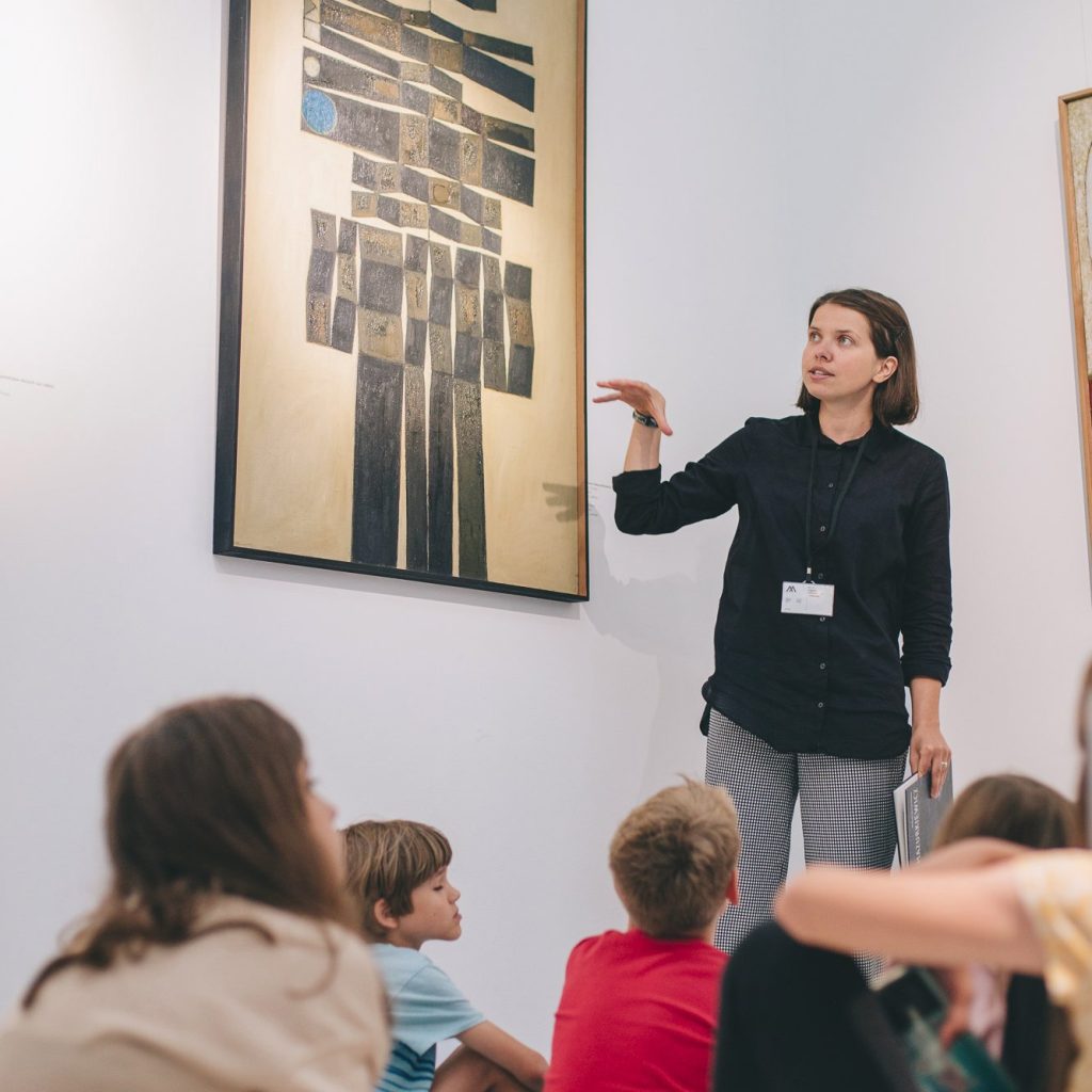 Kolorowa fotografia wykonana podczas wycieczki do muzeum. Młoda kobieta stoi przy obrazie i opowiada o nim, na podłodze siedzą zasłuchane dzieci.