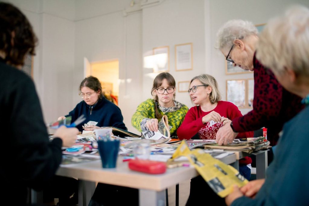 zdjęcie: Paweł Rychert Kolorowa fotografia wykonana podczas warsztatów plastycznych. W jasnej sali, przy dużym stole siedzą kobiety w różnym wieku, robią kolaże. Przed nimi na stole leżą wycinki gazet, archiwalne zdjęcia i przybory plastyczne.