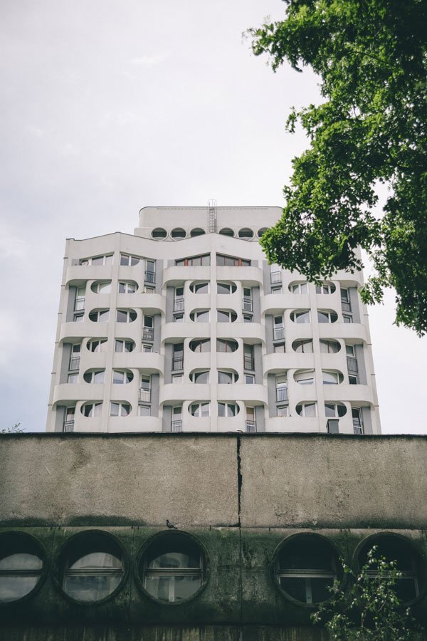 Widok na wysoki biały budynek z charakterystycznymi zaokrąglonymi oknami, tzw. Sedesowiec, na tle błękitnego lekko zachmurzonego nieba. 
