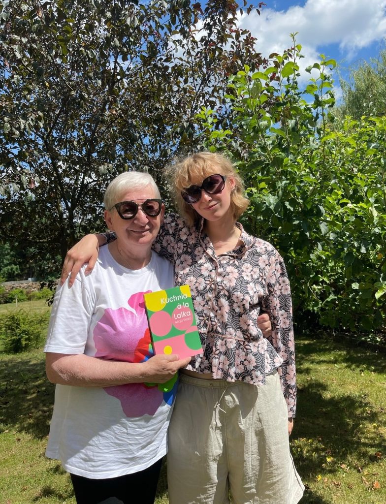 Kolorowa fotografia przedstawia seniorkę i młodą kobietę pozujące pod drzewami. Młodsz - Julia, obejmuję ręką starszą - swoją babcię, która w rękach trzyma książkę autorstwa wnuczki.