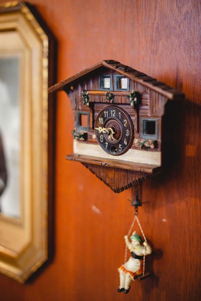 Zbliżenie na misternie wykonany zegar ścienny, przypominający wyglądaem ścianę drewnianej chaty. Funkcję wahadła pełni figurka dziewczynki, siedzącej na huśtawce.