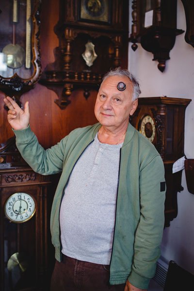 Portret pana Janusza, zegarmistrza. Starszy mężczyzna z charakterystyczną zegarmistrzowską lupą na czole pozuje do zdjęcia, wskazując zegary wiszące za nim na ścianie.