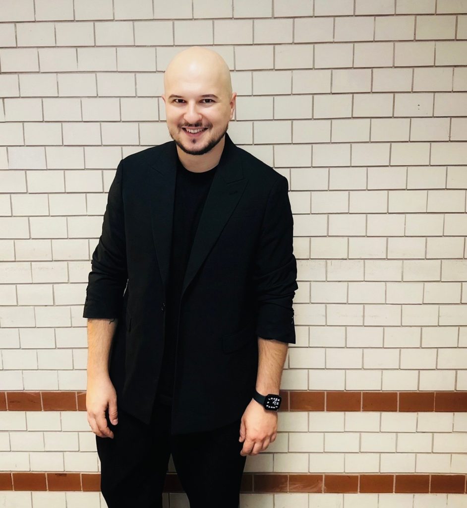 Kolorowa fotografia przedstawia łysego mężczyznę z brodą i pewnym siebie uśmiechem, stojącego przed dwubarwną ceglaną ścianą. Ubrany jest w czarną marynarkę, koszulkę i spodnie.