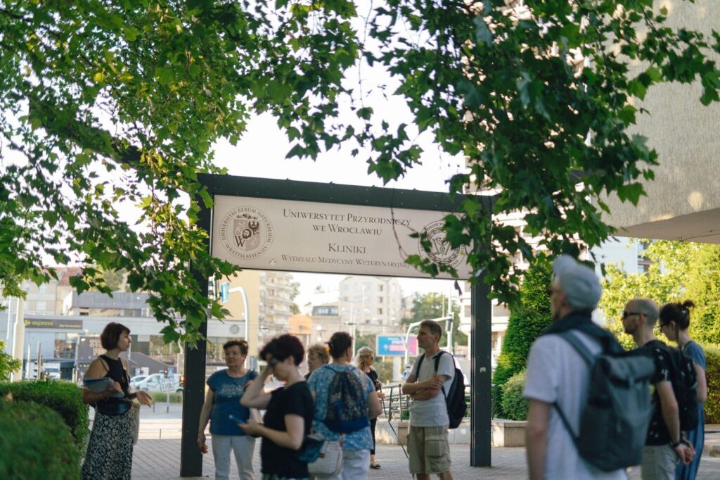 zdjęcie: Marcin Szczygieł Kolorowa fotografia przedstawia grupę osób zebranych wokół tablicy z napisem: Uniwersytet Przyrodniczy we Wrocławiu, Kliniki.