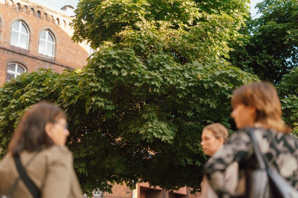 zdjęcie: Marcin Szczygieł Kolorowa fotografia przedstawia niewyraźne osoby na pierwszym planie, w tle za nimi potężne drzewo.