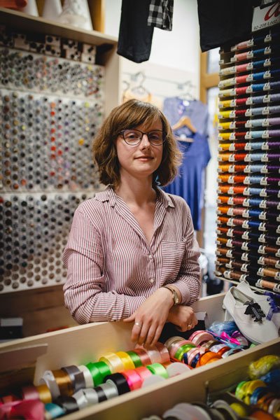 Pani Joanna, młoda kobieta w różowej koszuli w jasne prążki, zapinanej na guziki, stoi za ladą pasmanterii. Dookoła niej mnóstwo nici, guzików, tasiemek i zamków błyskawicznych.