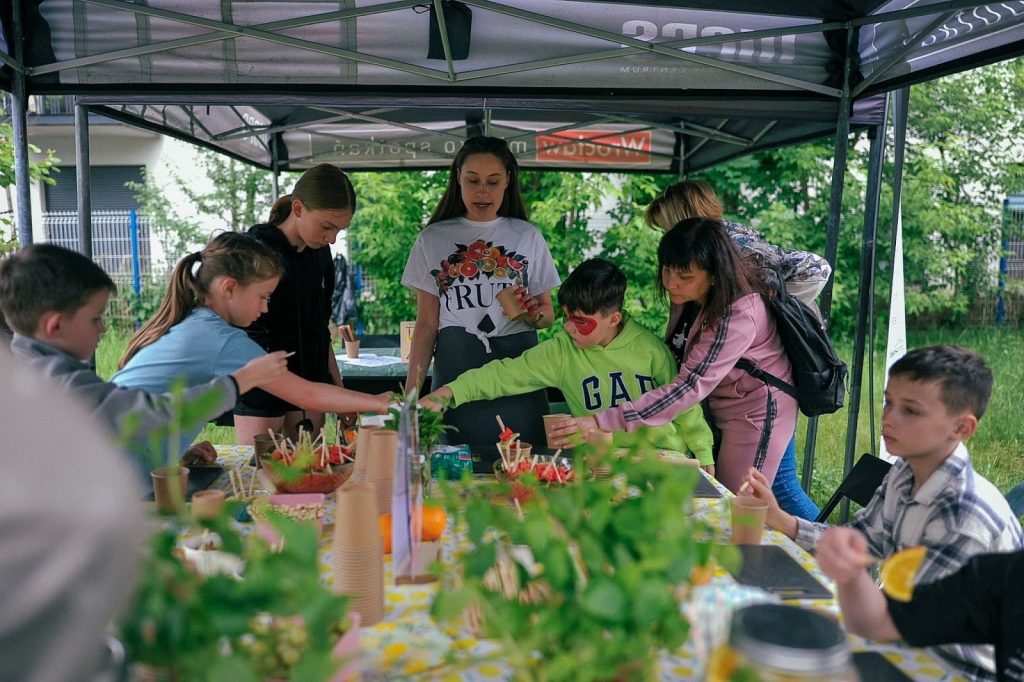 zdjęcie: Anna Pazdej Kolorowa fotografia wykonana podczas pikniku na osiedlu: pod dużym namiotem rozstawionym na łące trwają warsztaty dla dzieci. Grupa dzieci zebrała się wokół dużego stołu i prowadzącej. Na stole porozstawiane są owoce i zioła. Za chwilę wszyscy będą robić zdrowe lemoniady.