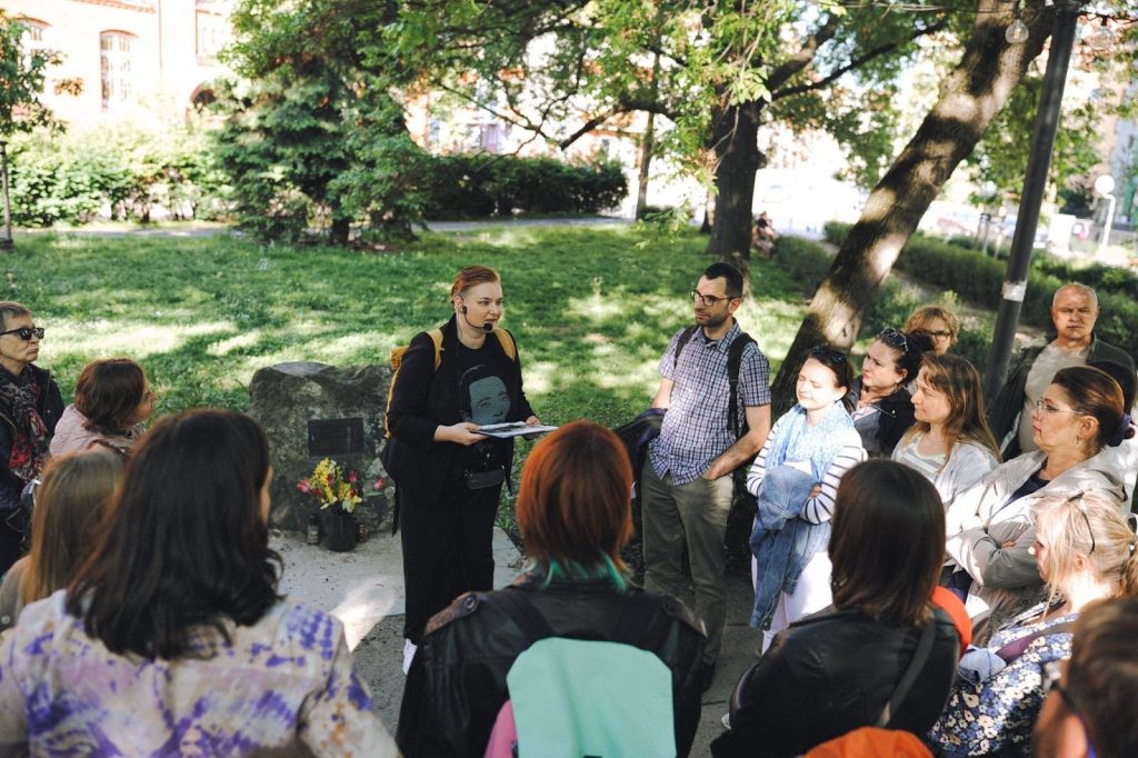 zdjęcie: Anna Pazdej Kolorowa fotografia wykonana podczas spaceru po osiedlu. Wokół przewodniczki zebrała się duża grupa osób w różnym wieku, która słucha o historii miejsca, w którym się znajdują.