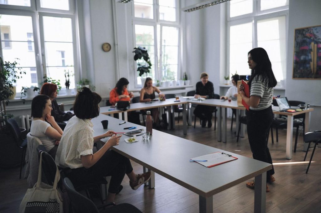 zdjęcie: Anna Pazdej Kolorowa fotografia wykonana podczas warsztatów. W dużym , jasnym pomieszczeniu przy długich stołach siedzą osoby w różnym wieku. Przed nimi stoi prowadząca spotkanie.