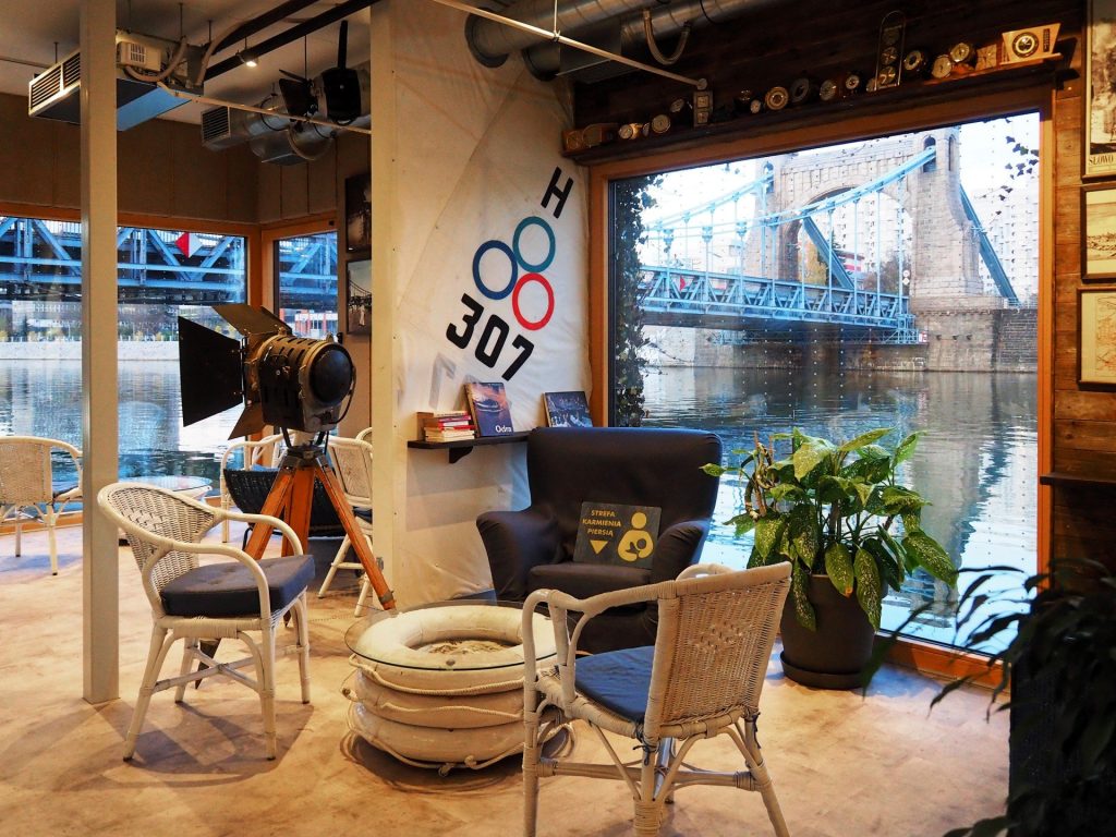 Wnętrze Odra Centrum. Przez duże okno widoczny most Grunwaldzki. Pod oknem ustawione są krzesła i stolik zrobiony z białych kół ratunkowych.