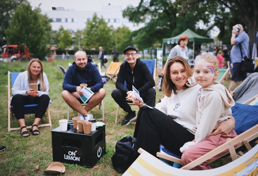 zdjęcie: Anna Pazdej Kolorowa fotografia wykonana podczas osiedlowego pikniku. Na leżakach rozstawionych na łące siedzą młodzi dorośli oraz dziewczynka z pomalowaną twarzą. Usmiechają się i patrzą w stronę aparatu.