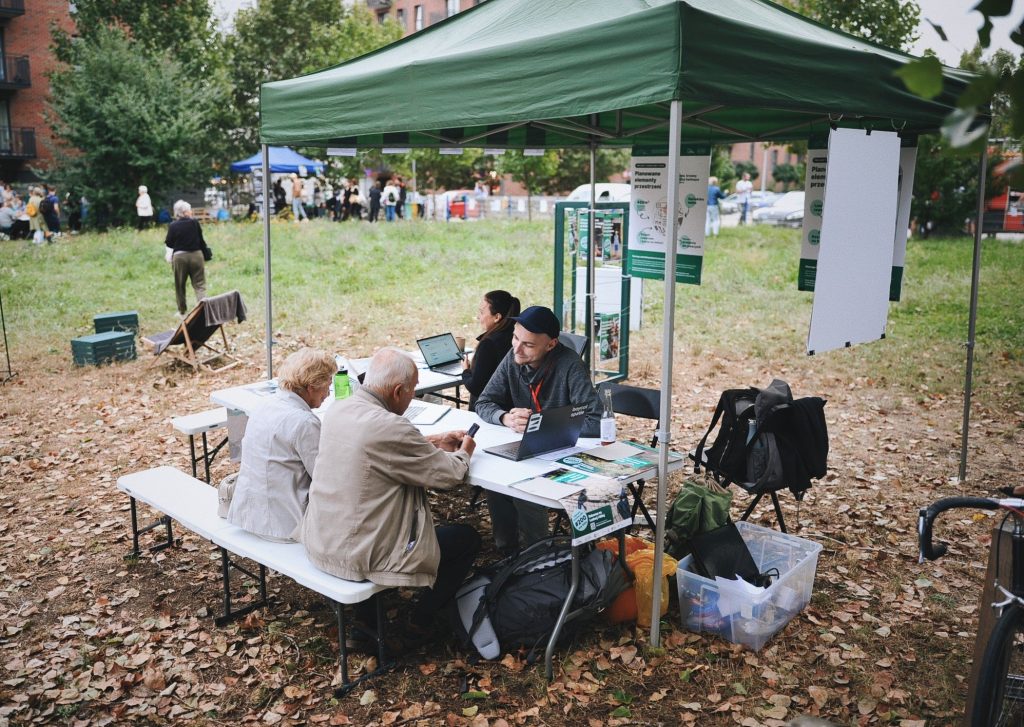 zdjęcie: Anna Pazdej Kolorowa fotografia wykonana podczas osiedlowego pikniku. Przy stoliku stojącym pod namiotem rozstawionym na łące siedzi dwójka seniorów oraz młody mezczyzna opowiadający o WBO.