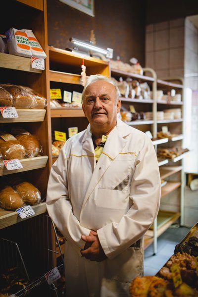 Pan Ryszard, starszy mężczyzna w białym fartuchu, na tle półek z pieczywem.