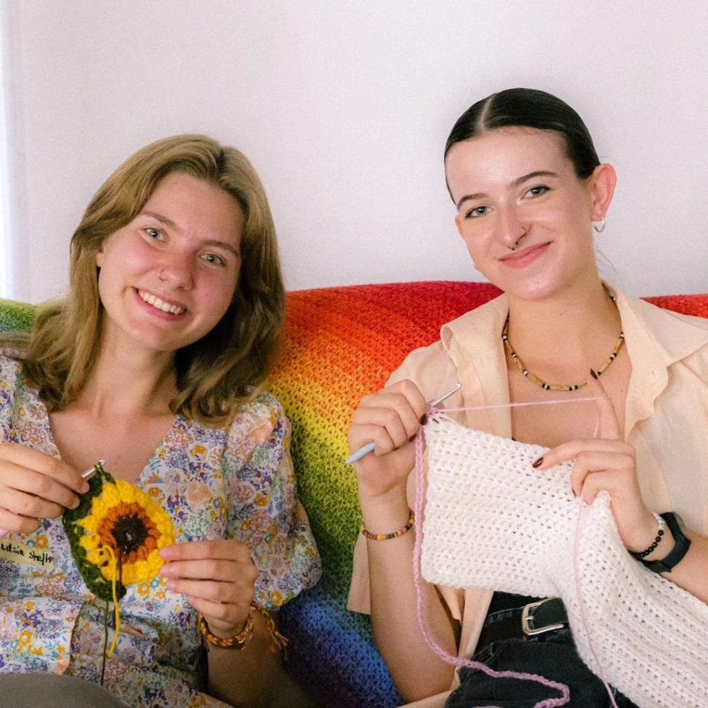 Kolorowa fotografia przedstawia dwie uśmiechnięte osoby robiące na szydełku.