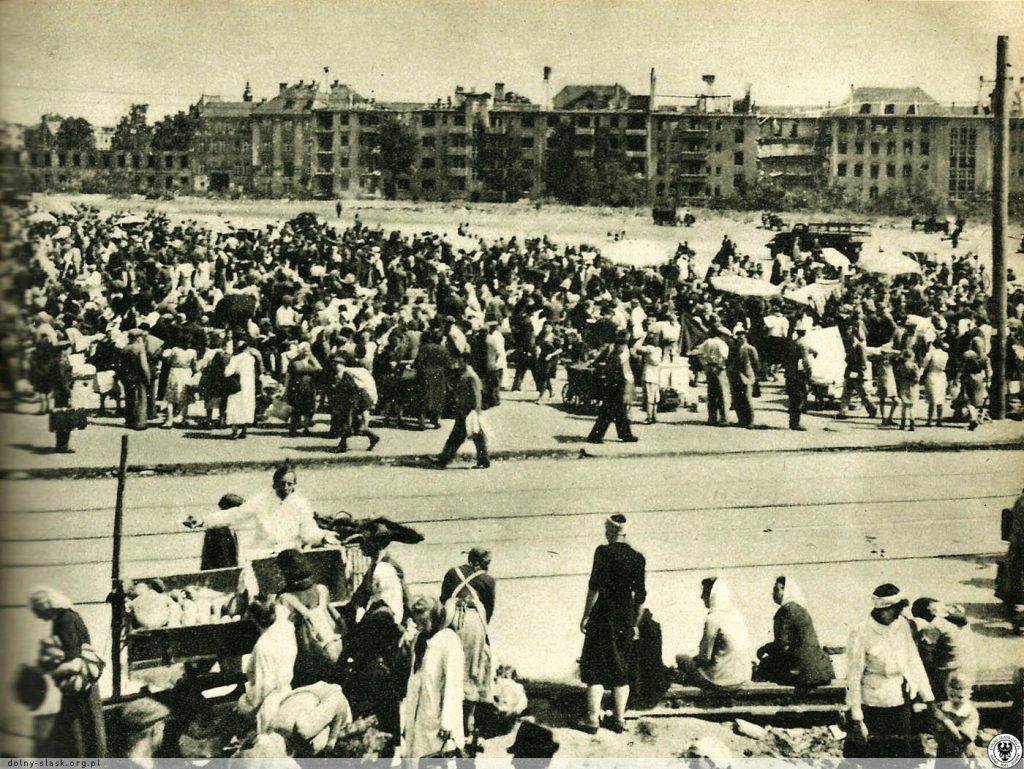 Szaberplac na pl. Grunwaldzkim. Na pierwszym planie torowisko na ul. Piastowskiej, a w tle Akademia Rolnicza i zabudowa ul. Norwida lata 1945-1948. Fot. za Wratislaviae Amici (https://polska-org.pl/856347,foto.html?idEntity=510862) Archiwalne zdjęcie w sepii: szaberplac na placu Grunwaldzkim. Dużo ludzi tłoczy się po dwóch stronach ulicy, oddzielonych torowiskiem. W tle zniszczona zabudowa ul. Norwida.