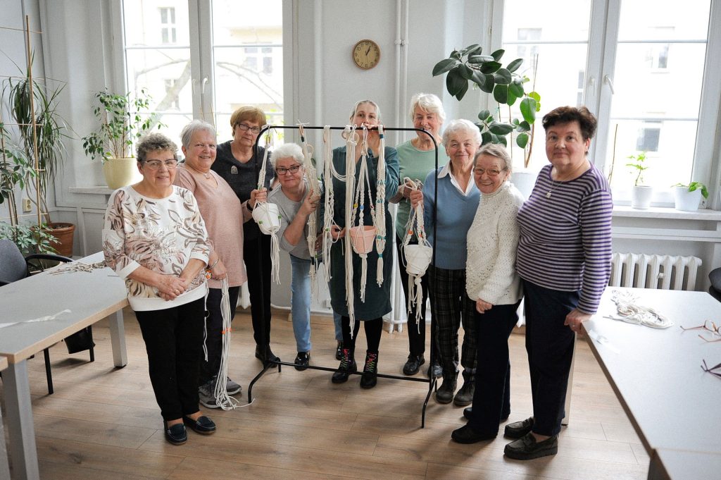 zdjęcie: Anna Pazdej Kolorowa fotografia przedstawia grupę seniorek pozujących do zdjęcia z wyplecionymi na zajęciach kwietnikami.