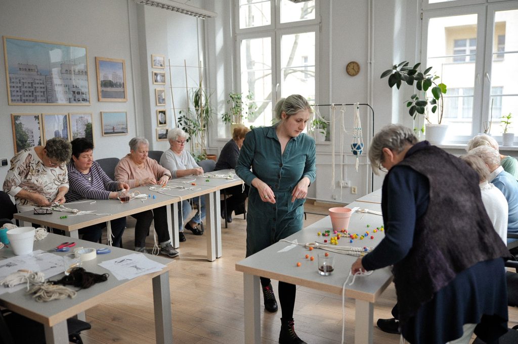 zdjęcie: Anna Pazdej Kolorowa fotografia wykonana podczas warsztatów rzemieślniczych dla seniorów i seniorek. W jasnym pomieszczeniu przy długich stołach siedzą seniorki i wyplatają ze sznurków kwietniki. Między stołami chodzi młoda kobieta prowadząca spotkanie.