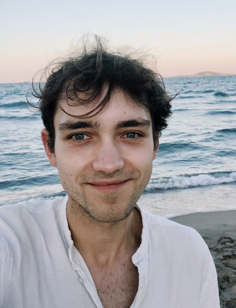 Kolorowa fotografia przedstawia uśmiechniętego młodego mężczyznę na tle morza.