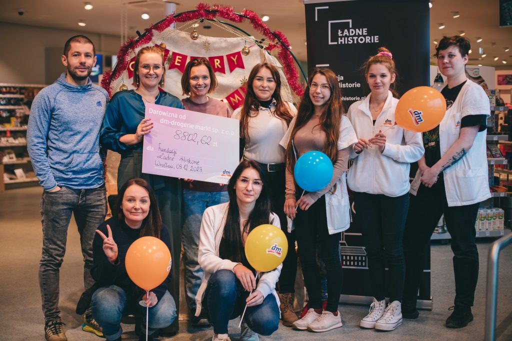 zdjęcie: Marta Sobala Kolorowa, pozioma fotografia: zespół Ładnych Historii oraz pracowniczki drogerii dm pozują do zdjęcia na tle sklepu i baneru fundacji.