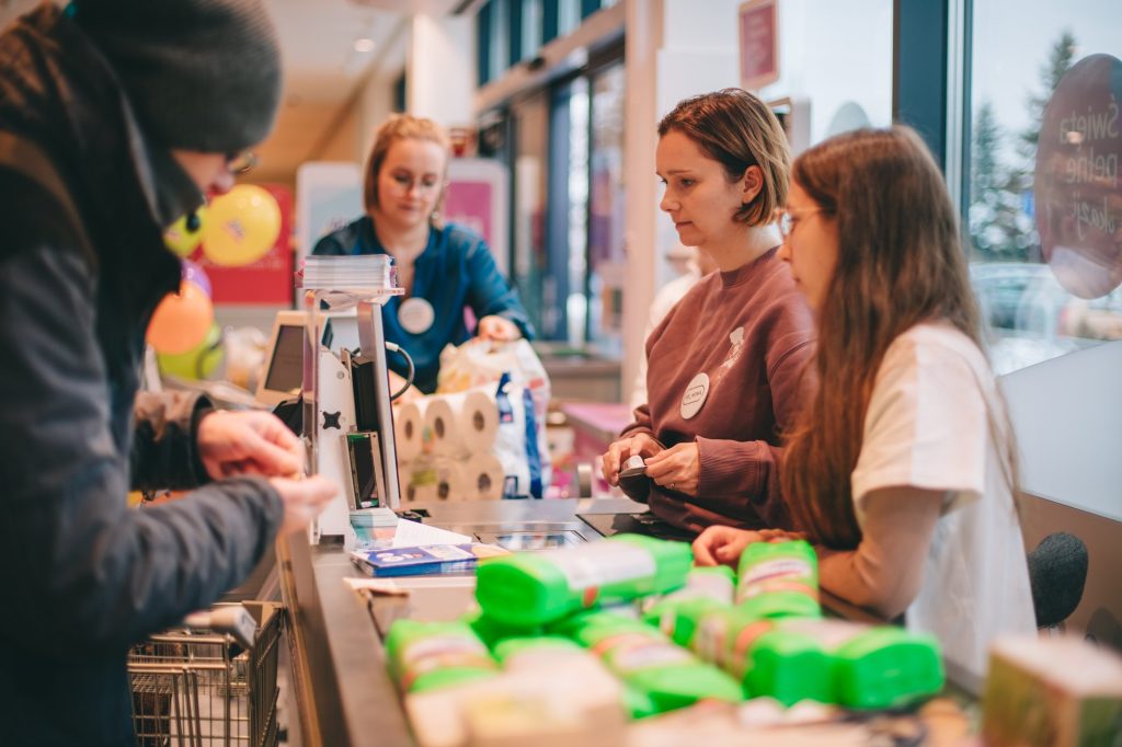 zdjęcie: Marta Sobala Kolorowa, pozioma fotografia: przy kasie w drogerii dm siedzi Kornelia z Fundacji Ładne Historie i kasuje zakupy w ramach akcji charytatywnej dla seniorów i seniorek. W tle widoczna również Ania z Fundacji, która pakuje zakupy.