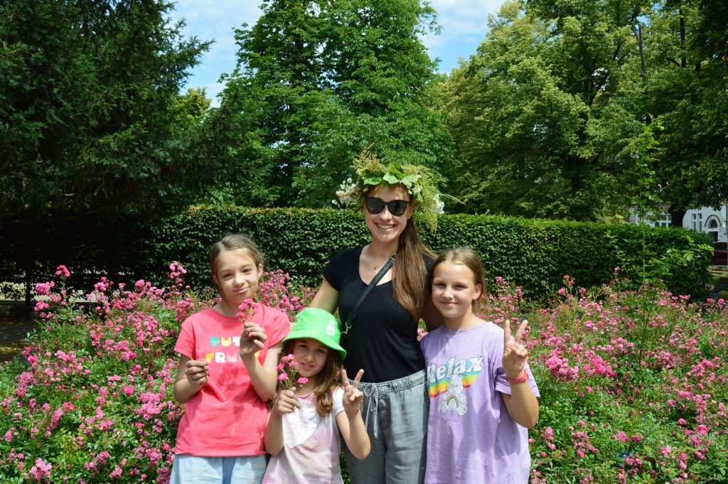 Kolorowa fotografia: młoda kobieta z kolorowym wiankiem na głowie pozuje do zdjęcia razem z trzema młodymi dziewczynkami na tle bujnego, kolorowo kwitnącego krzaka. Kolorowa fotografia: młoda kobieta z kolorowym wiankiem na głowie pozuje do zdjęcia razem z trzema młodymi dziewczynkami na tle bujnego, kolorowo kwitnącego krzaka.