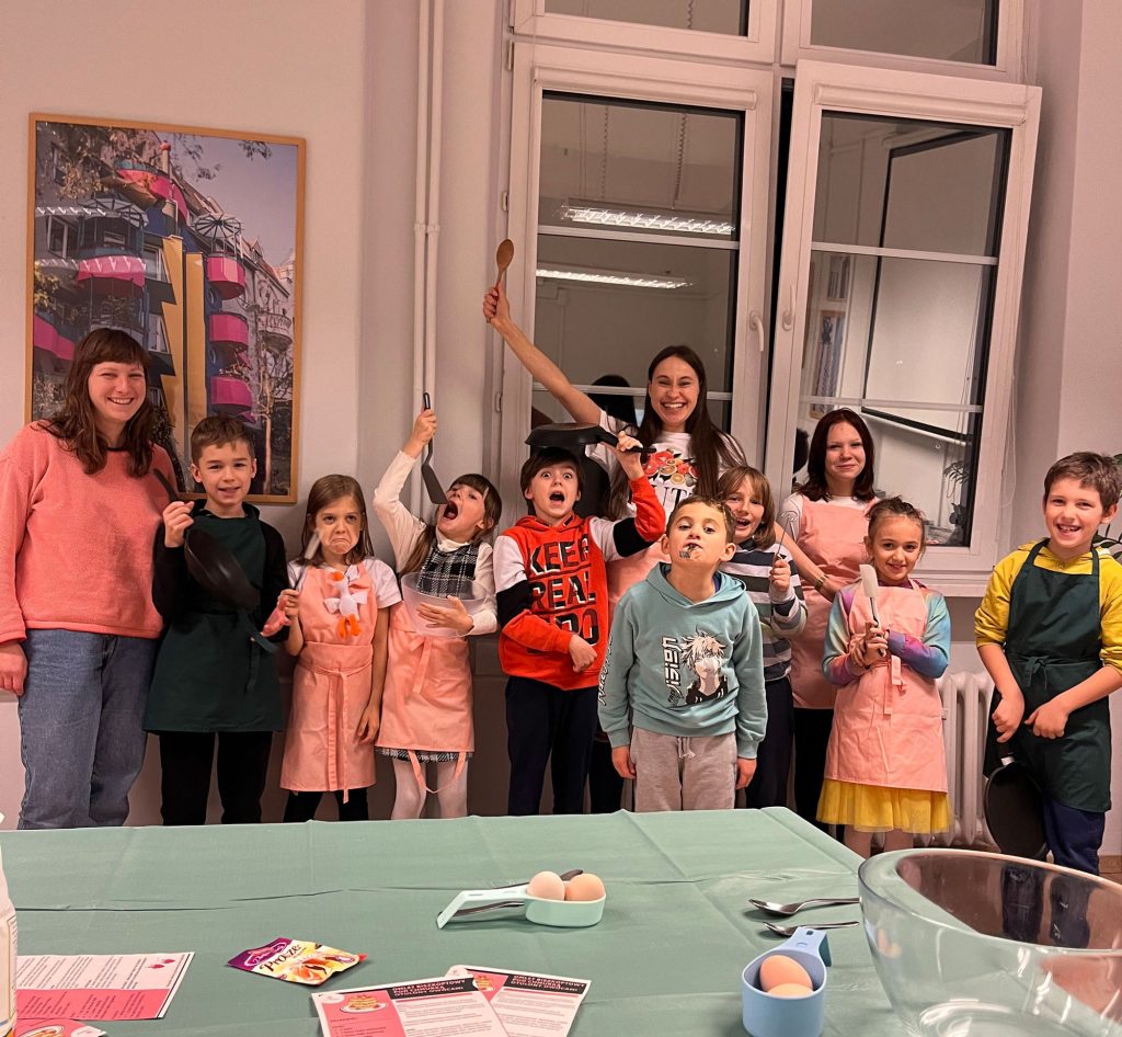 Kolorowa, pozioma fotografia: grupka dzieci w różnym wieku oraz dwie animatorki pozują do zdjęcia w jasnym pomieszczeniu, po zakończonych warsztatach kulinarnych. Dzieciaki machają trzymanymi w rękach łyżkami, chochlami i lizawkami. Na ich twarzach widać radość. Kolorowa, pozioma fotografia: grupka dzieci w różnym wieku oraz dwie animatorki pozują do zdjęcia w jasnym pomieszczeniu, po zakończonych warsztatach kulinarnych. Dzieciaki machają trzymanymi w rękach łyżkami, chochlami i lizawkami. Na ich twarzach widać radość.