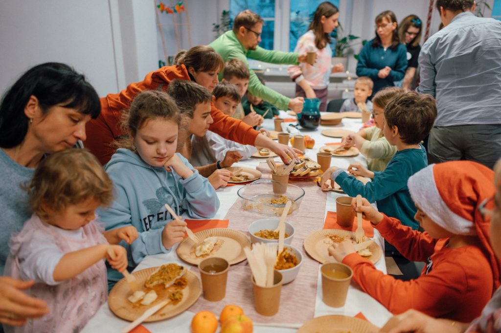 zdjęcie: Marcin Szczygieł Kolorowa, pozioma fotografia: w pomieszczeniu przy długim stole siedzi duża grupa osób w różnym wieku, trwa osiedlowe spotkanie świąteczne. Wszyscy jedzą wspólnie przygotowane smakołyki, rozmawiają i śmieją się.